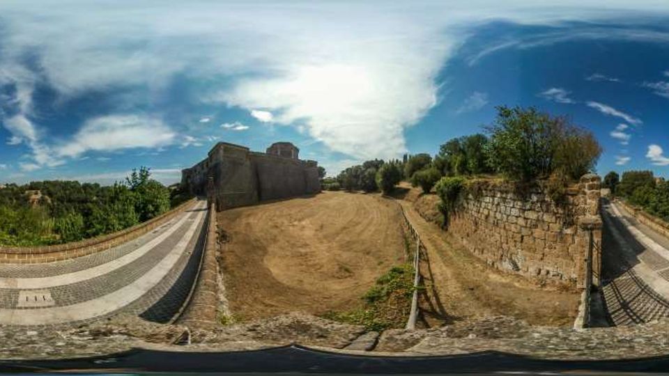Vista panoramica di un’area all’aperto con un edificio in pietra di tipo fortificato e un ampio spiazzo sterrato centrale. Ai lati si vedono rampe pavimentate con cordoli in pietra e un alto muro in blocchi di pietra, con vegetazione e cielo parzialmente nuvoloso sullo sfondo.