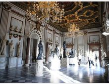 Ampia sala museale in stile neoclassico con pavimento a scacchi, pareti ornate da nicchie con statue e busti, e grandi lampadari di cristallo sospesi sotto un soffitto riccamente decorato. Al centro della stanza sono esposte sculture su piedistalli, con luce naturale che entra da ampie porte e finestre.
