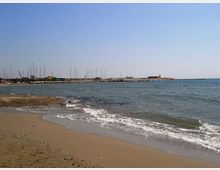 Spiaggia sabbiosa con piccole onde in primo piano, affacciata su un molo frangiflutti e un porto con numerosi alberi di barche a vela sullo sfondo. Cielo sereno e mare aperto a destra.
