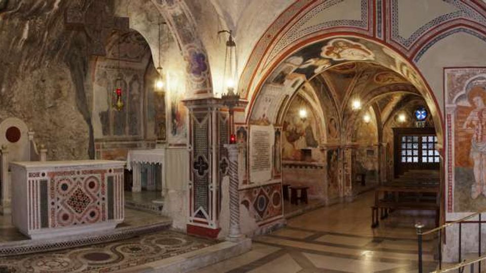 Interno di una chiesa o cappella sotterranea con navate ad archi e volte affrescate, pareti in parte scavate nella roccia e pavimento a mosaico. In primo piano si vede un altare in pietra e, sul fondo, una porta finestrata con panche lungo il corridoio.