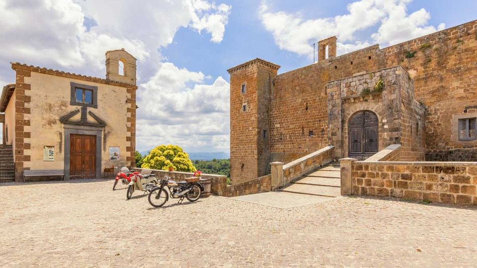 Piazza lastricata in pietra con una piccola chiesa con campanile a vela e, a destra, un grande edificio fortificato in muratura con torre e portale ad arco raggiunto da una rampa con parapetti. Sullo sfondo si vedono alberi e colline sotto un cielo azzurro con nuvole.