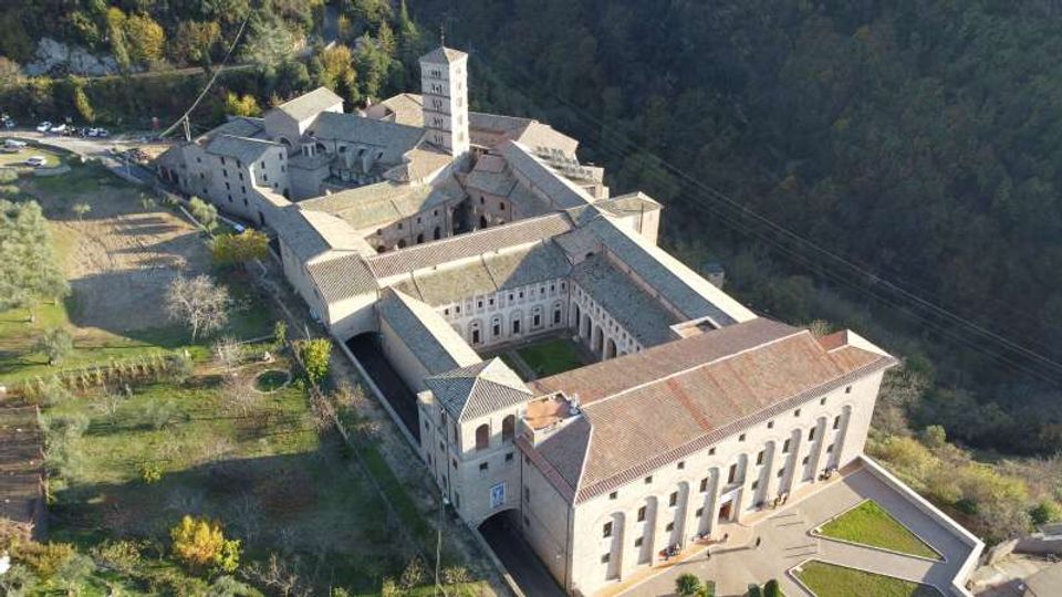 Veduta aerea di un grande complesso monastico in pietra con tetti in coppi, organizzato attorno a chiostri e cortili interni, con un campanile quadrato centrale. L’edificio è circondato da aree verdi e si trova ai margini di un bosco su un pendio.