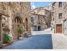 Strada acciottolata in un borgo storico, fiancheggiata da edifici in pietra con archi, finestre piccole e porte in legno. La via curva e sale leggermente tra facciate rustiche con vasi di piante e segnaletica stradale.