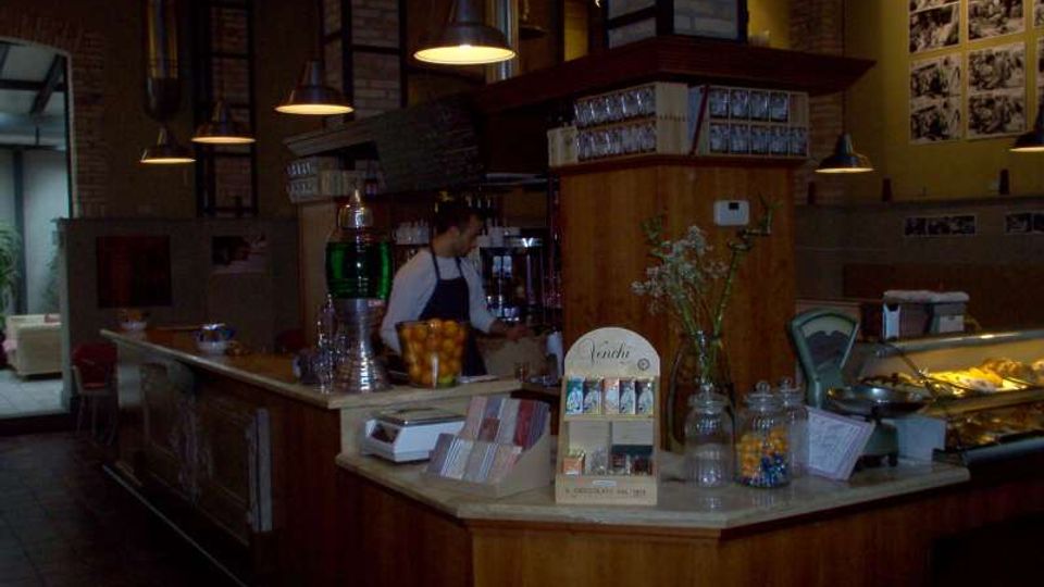 Interno di un bar-caffetteria con bancone in legno e vetrina espositiva, illuminato da lampade a sospensione. Pareti in mattoni a vista e pannelli fotografici alle pareti, con area di servizio e attrezzature dietro il bancone.