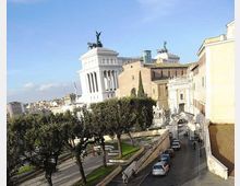 Veduta urbana con un grande monumento in marmo bianco a colonne, sormontato da statue equestri e una quadriga, sullo sfondo. In primo piano si vede una strada in discesa con auto parcheggiate, marciapiedi e alberi lungo un’area verde, affiancata da edifici storici.
