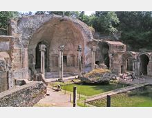 Rovine di un complesso termale romano con grandi volte in muratura, colonne in pietra e archi laterali. In primo piano si vedono vasche rettangolari con acqua verdastra e un grande masso, circondate da percorsi e aree recintate, con alberi sullo sfondo.
