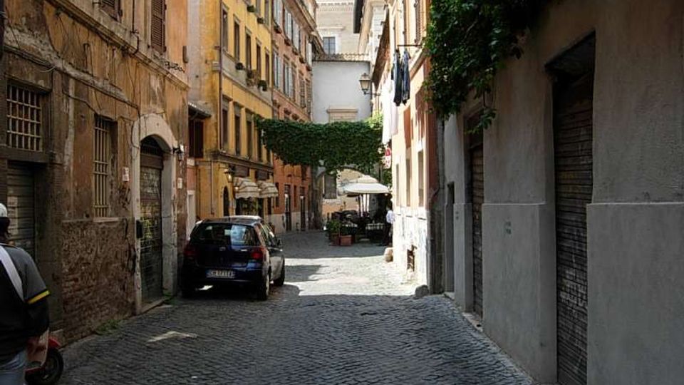 Strada stretta in acciottolato tra alti edifici storici con facciate color ocra e persiane, con un’auto parcheggiata sul lato sinistro. In fondo si vede una piccola piazzetta ombreggiata con alberi e tavolini sotto ombrelloni.