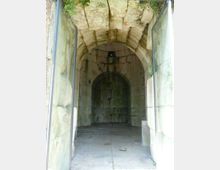 Interno di un passaggio in pietra con volta a botte e pareti di grandi blocchi, che conduce a una nicchia ad arco sul fondo. Ai lati si vedono pannelli di vetro incorniciati e sul pavimento lastre di pietra con piccole chiazze di vegetazione.