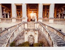 Cortile interno di un palazzo storico con doppia scalinata in pietra e balaustre, che converge verso una terrazza centrale. Al livello superiore si vedono colonne, nicchie con statue e un portale illuminato sullo sfondo.