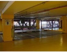 Interno di un parcheggio multipiano con pilastri e soffitto gialli, tubazioni e lampade a vista, e posti auto delimitati da strisce bianche. Sul pilastro è presente un cartello “Parcheggio A Livello -1” con il numero 9 e sul fondo si vedono pannelli a griglia che lasciano filtrare la luce dall’esterno.
