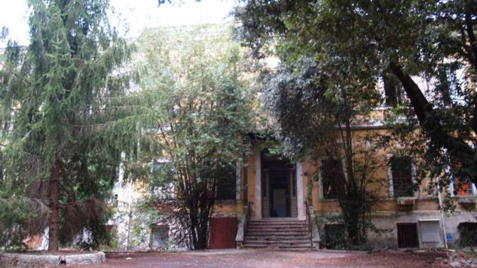 Ingresso di un edificio a due piani con facciata chiara, raggiunto da una breve scalinata centrale. L’area antistante è un cortile in terra battuta circondato da alberi alti e vegetazione fitta che ombreggia la struttura.
