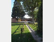 Vialetto pavimentato che attraversa un prato e conduce a un complesso di edifici bassi con porticati in legno, delimitato da staccionate. Grandi alberi ad alto fusto e vegetazione fitta incorniciano l’area esterna.