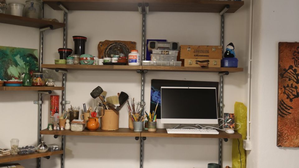 Angolo di studio/laboratorio artistico con pareti bianche e scaffalature in legno e metallo piene di barattoli, colori, pennelli e materiali da lavoro. Una lampada a sospensione illumina un ripiano con un monitor e, sotto, contenitori e scatole organizzate su pavimento in legno.