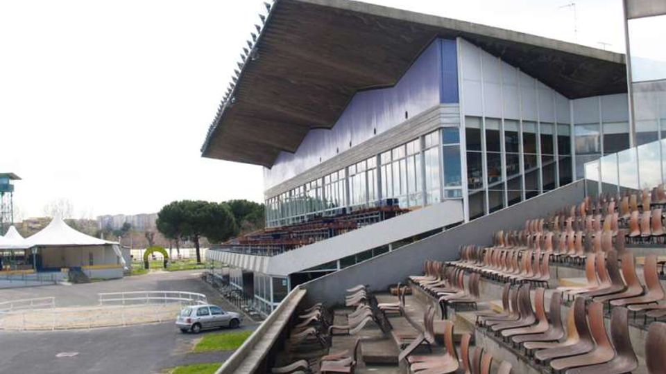Tribuna di uno stadio o ippodromo con gradinate in cemento e file di sedili in plastica marrone, affiancata da un edificio con ampie vetrate e grande tetto sporgente. In basso si vedono l’area esterna con strada, parcheggio e alcune strutture temporanee.