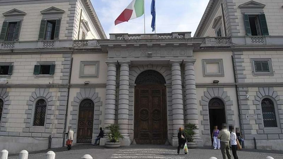 Facciata di un edificio istituzionale in stile neoclassico, con ingresso centrale ad arco affiancato da colonne e grandi portoni in legno. In cima sventolano le bandiere italiana e dell’Unione Europea, davanti a un piazzale lastricato con dissuasori in pietra.