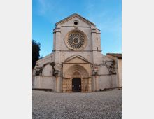 Facciata di una chiesa in pietra con grande rosone circolare centrale e portale ad arco ogivale, affiancata da contrafforti e arcate laterali. Davanti si vede una piazza lastricata in pietra sotto un cielo azzurro.