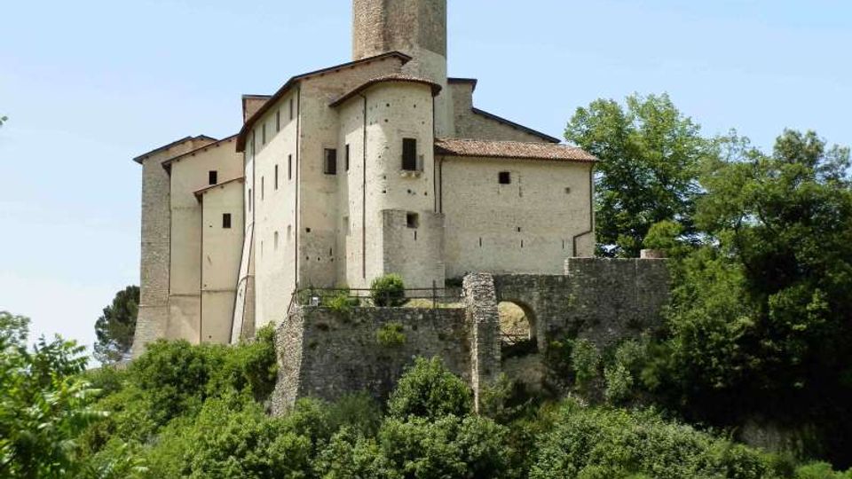 Castello in muratura chiara su una collina, con torre cilindrica centrale e tetti in coppi rossi. In primo piano si vede un muraglione di pietra con arco e vegetazione fitta tutt’intorno.