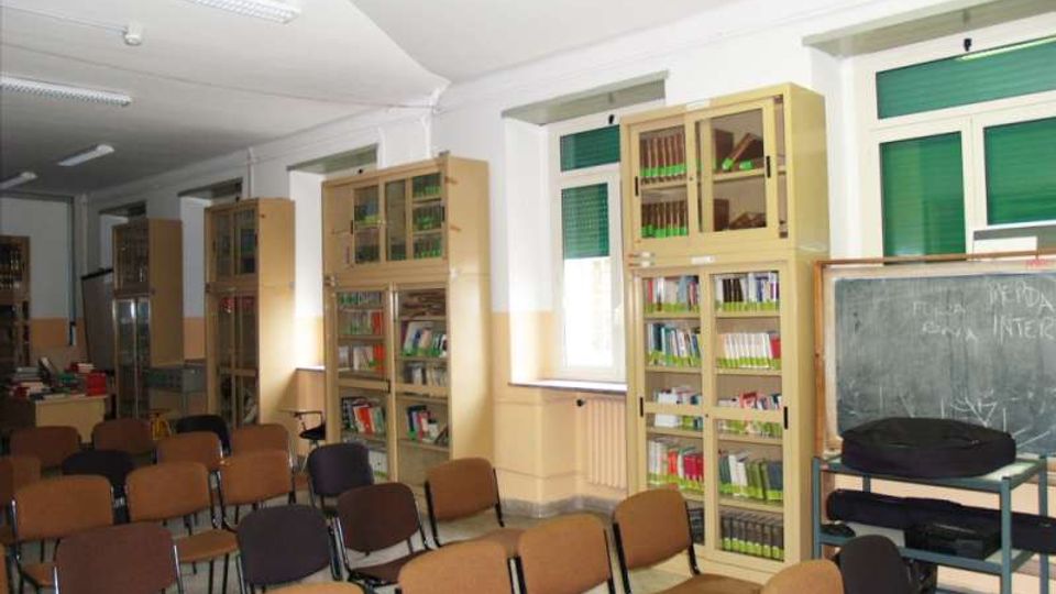 Aula scolastica con file di sedie rivolte verso una lavagna su carrello, pavimento chiaro e illuminazione a neon sul soffitto. Lungo le pareti ci sono alte librerie con ante in vetro e finestre con tapparelle verdi.
