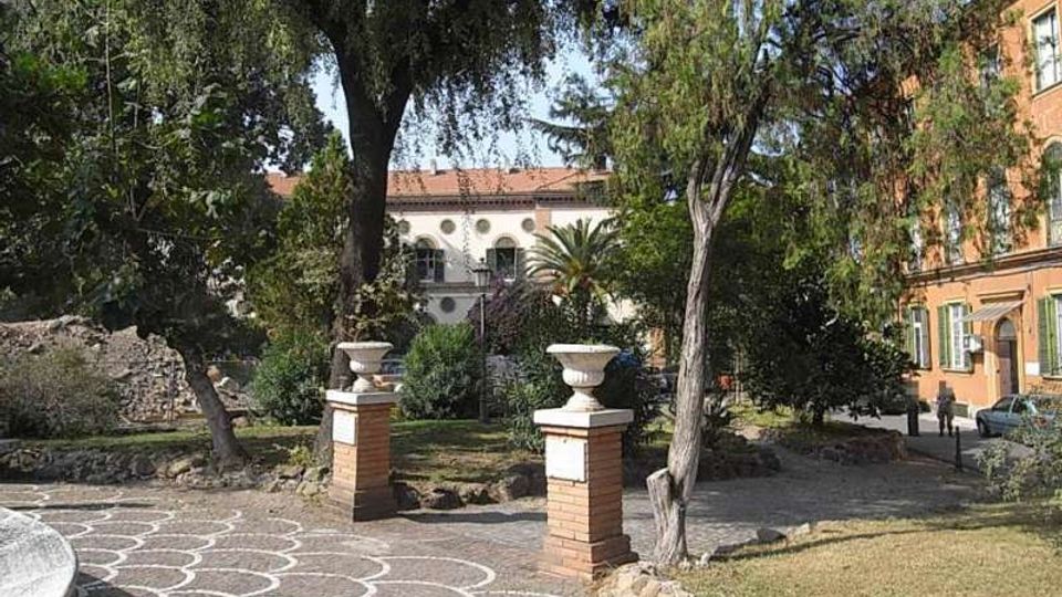 Piccolo parco urbano con vialetto in pietra decorato a motivi circolari bianchi e due pilastri in mattoni con vasi all’ingresso. L’area è circondata da alberi e aiuole, con edifici storici sullo sfondo e una strada laterale.