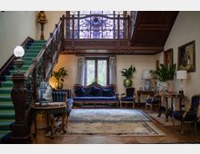 Interno di un salone in stile classico con grande scala in legno e ringhiera in ferro battuto, soppalco con vetrata decorata e pavimento in parquet con ampi tappeti. Divani e poltrone blu, tavolini con lampade e vasi, piante in vaso e quadri alle pareti vicino a una portafinestra con tende.