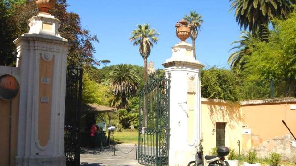Ingresso di un parco o giardino con due alti pilastri in muratura chiara sormontati da vasi ornamentali e un cancello in ferro battuto aperto. All’interno si vedono vialetti e vegetazione con alte palme, mentre a destra corre un muro color ocra lungo la strada.