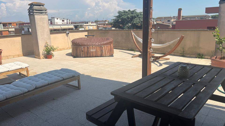 Terrazza sul tetto pavimentata con parapetto basso, arredata con tavolo e panche in legno, lettini, una vasca idromassaggio rotonda e un’amaca su supporto in legno. Sullo sfondo si vede il panorama dei tetti e degli edifici circostanti sotto un cielo sereno con grandi nuvole.
