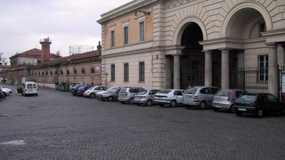 Strada lastricata in pietra con auto parcheggiate lungo il marciapiede, affiancata da un grande edificio storico con facciata in pietra e ampio portico ad archi sostenuto da colonne. Sullo sfondo si estende un lungo muro in mattoni con finestre ad arco e tetti bassi.