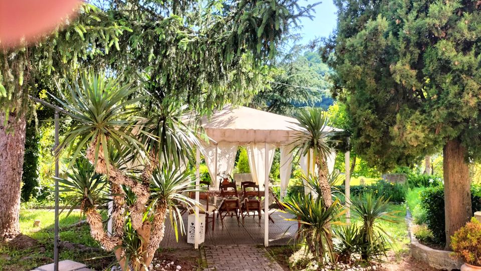 Gazebo da giardino con tetto beige e tende bianche, con tavoli e sedie su un vialetto in pietra. L’area è circondata da alberi alti e piante ornamentali, con aiuole e prato in un contesto verde all’aperto.