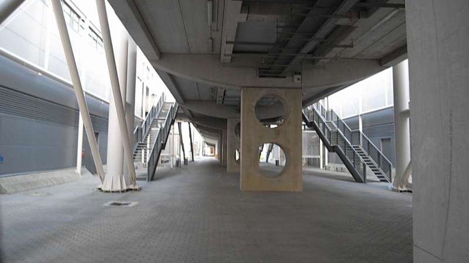 Passaggio pedonale coperto sotto una struttura sopraelevata in cemento e acciaio, con pavimentazione in blocchetti e colonne portanti inclinate. Ai lati si vedono scale metalliche e, al centro, un setto in cemento con aperture circolari, circondato da edifici industriali moderni.