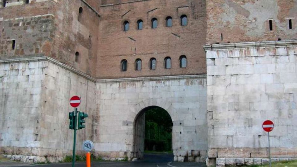Ingresso ad arco in una cinta muraria di un edificio storico in mattoni con merli e piccole finestre, poggiato su una base in pietra chiara. Davanti passa una strada asfaltata con segnaletica di divieto e un semaforo.