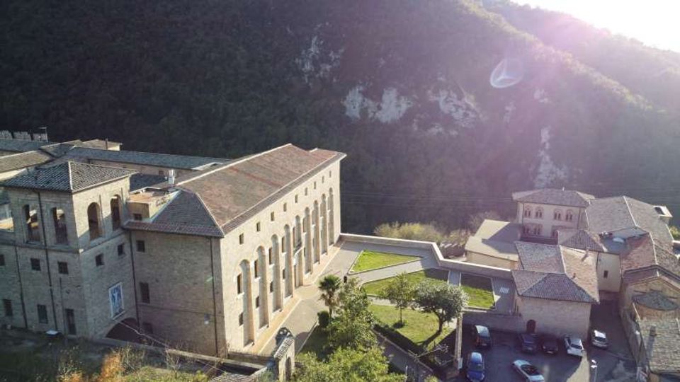 Complesso di edifici in pietra con tetti in tegole, dominato da un grande edificio rettangolare con alte finestre ad arco, affacciato su un cortile e aree verdi. Sullo sfondo si vede un pendio montuoso boscoso, con strada e piccoli edifici adiacenti.