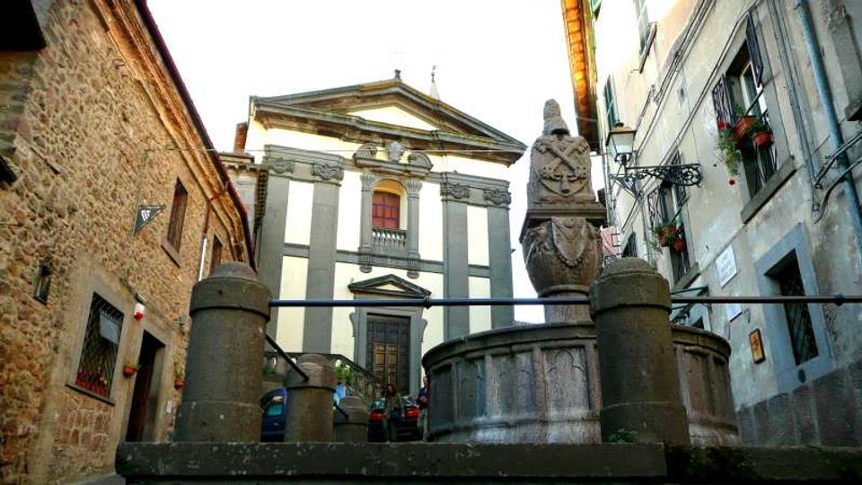 Piccola piazza tra edifici in pietra e intonaco, con una fontana circolare in primo piano sormontata da una scultura. Sullo sfondo si vede la facciata di una chiesa con frontone e lesene, raggiungibile tramite una breve scalinata.