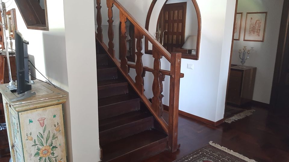 Interno di una casa con scala in legno scuro e corrimano intagliato che sale lungo una parete bianca. Sul pianerottolo si vede una grande specchiera ad arco, pavimento in parquet con tappeto e mobili in legno, tra cui una cassettiera dipinta e una piccola zona con quadri alle pareti.
