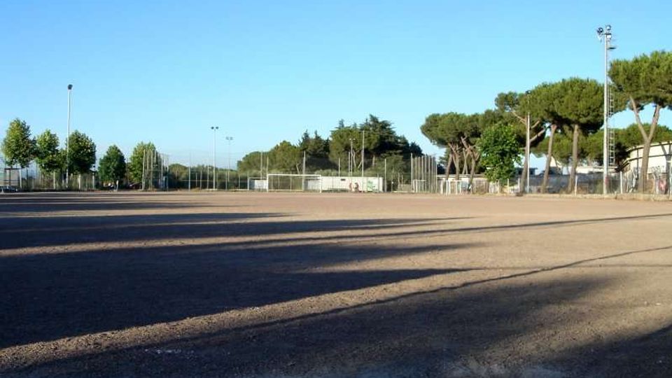 Ampio campo sportivo in terra battuta recintato, con un cancello e pali dell’illuminazione; sullo sfondo si vedono alberi ad alto fusto e alcune strutture basse sotto un cielo sereno.