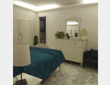 Camera da letto luminosa con pareti bianche, pavimento in marmo chiaro e letto con copriletto blu. Sono visibili una cassettiera bianca con grande specchio decorato, armadio, lampade da tavolo e una porta aperta verso un altro ambiente.