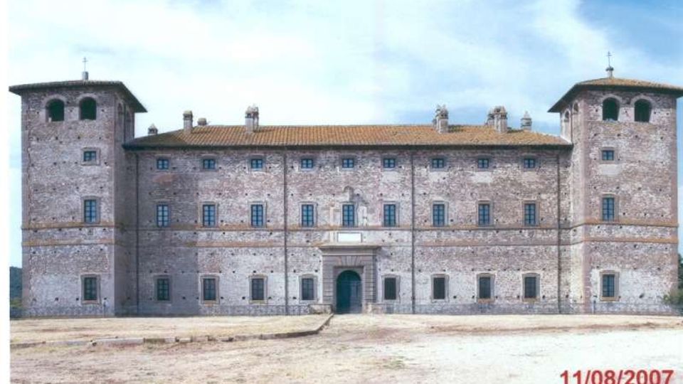 Facciata di un grande castello in mattoni con due torri quadrate laterali, tetto in coppi e file regolari di finestre. Al centro si vede un ingresso ad arco con portale, davanti a un ampio spiazzo in terra battuta sotto un cielo parzialmente nuvoloso.