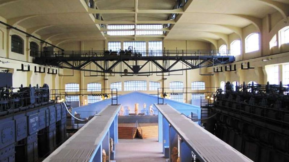 Interno di un grande edificio industriale riconvertito, con alti soffitti a volta, ampie finestre ad arco e una passerella metallica sospesa tra le pareti. Al centro si vede una lunga navata con due corridoi sopraelevati e strutture macchinari scuri ai lati.