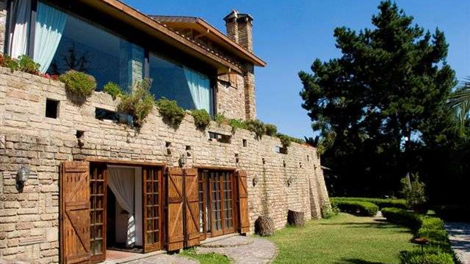 Casa in pietra a due piani con grandi finestre e portefinestre con persiane in legno, balconata con vasi di piante e tetto spiovente con camino. Davanti c’è un giardino curato con prato, siepi e vialetto in pietra, con alti alberi sullo sfondo.