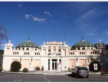 Facciata simmetrica di un grand hotel in stile classico, con due grandi cupole verdi laterali e ingresso centrale decorato con frontone e colonne. Davanti si vede un piazzale con posti auto, aiuole e lampioni, sotto un cielo sereno.