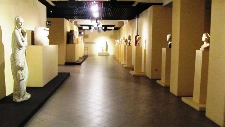 Corridoio interno di un museo o galleria con pavimento in piastrelle e illuminazione a soffitto, fiancheggiato da piedistalli che espongono sculture e busti. Le pareti chiare e la prospettiva lunga conducono verso un’area espositiva sul fondo.