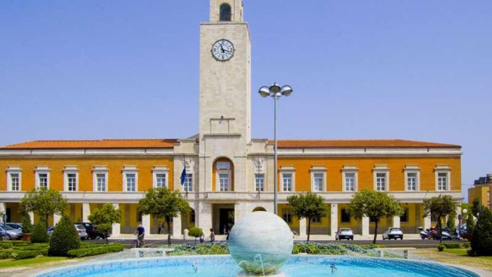 Edificio pubblico simmetrico in stile razionalista con facciata giallo ocra e una torre centrale in pietra con orologio. In primo piano si vede una grande fontana circolare con una sfera al centro, circondata da aiuole e alberi in una piazza.