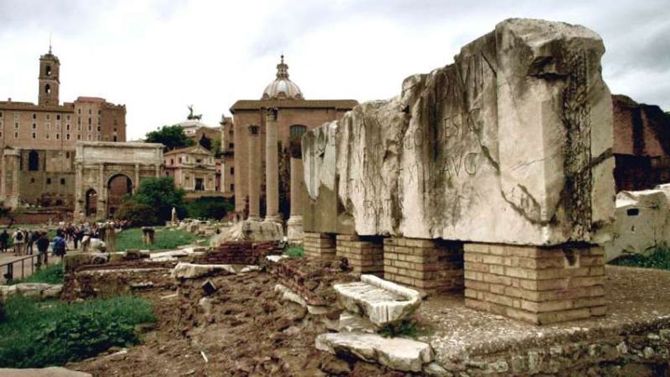 Veduta di un’area archeologica con grandi blocchi di marmo incisi su basamenti in mattoni e resti di muri e pavimentazioni in primo piano. Sullo sfondo si vedono edifici storici in muratura, un arco monumentale e una chiesa con cupola sotto un cielo nuvoloso.