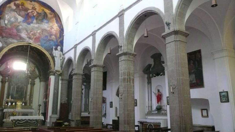 Interno di una chiesa con navata centrale, file di panche in legno e arcate in pietra su colonne che delimitano le navate laterali. Sul fondo si vede l’altare con un grande affresco nell’abside e alcune statue e quadri lungo le pareti bianche.