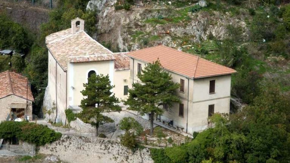 Complesso di edificio in muratura con piccola chiesa con campanile e tetti in coppi, situato su un pendio roccioso. Intorno si vedono alberi e vegetazione fitta, con pareti di roccia e una piccola terrazza murata davanti agli edifici.