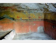 Interno di una tomba ipogea scavata nella roccia, con pareti dipinte da affreschi in giallo e rosso con fasce decorative e figure stilizzate. Sul lato sinistro si vede una banchina in pietra e il soffitto roccioso grezzo sopra la camera.