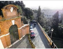 Strada in sampietrini in discesa accanto a un alto muro, con auto parcheggiate lungo il lato e lampione in primo piano. A sinistra si vede un grande portale in mattoni con arco e cancello in ferro, circondato da alberi e con vista su edifici sullo sfondo.