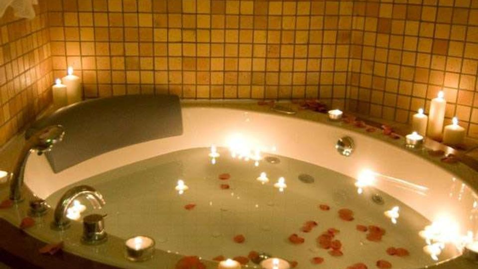 Vasca da bagno rotonda incassata in un bagno rivestito di piastrelle beige, con rubinetteria cromata e acqua. Candele accese e petali di rosa sono disposti sul bordo e sulla superficie dell’acqua, con asciugamani appesi sulla parete.