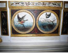 Dettaglio di una decorazione pittorica su soffitto con cornici dorate, composta da due medaglioni circolari affrescati che raffigurano uccelli in un paesaggio. Intorno sono presenti fasce ornamentali con motivi floreali e pannelli laterali scuri decorati.