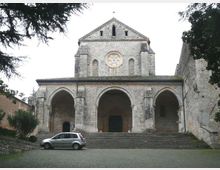 Facciata di una chiesa in pietra con ampio portico a tre arcate e lunga scalinata frontale; sopra il portico si vede un rosone centrale e piccole finestre ad arco. Davanti c’è una piazza lastricata, con mura laterali in pietra e alberi che incorniciano la scena.
