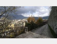 Strada acciottolata in pendenza con binari al centro, affiancata da una lunga muraglia in pietra a destra e da una ringhiera con alberi e case del paese a sinistra. Sullo sfondo si vedono montagne sotto un cielo nuvoloso.